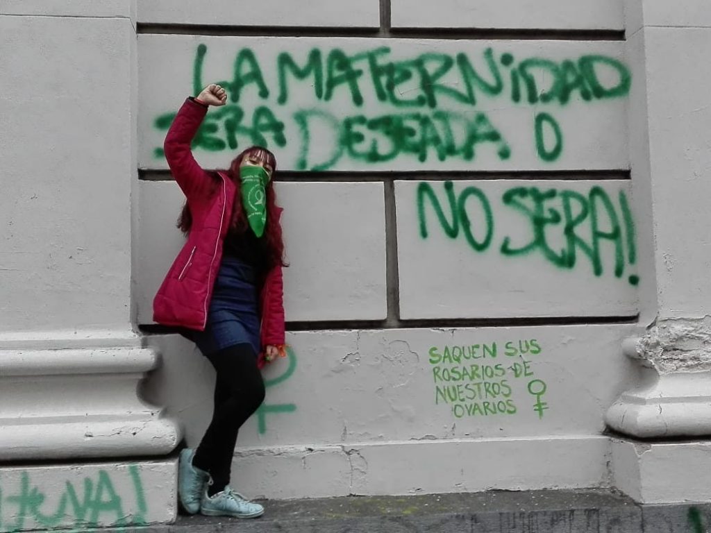 Привърженичка на легализацията на аборта пред надпис на стена в Буенос Айрес, който гласи: "Майчинството или трябва да бъде желано, или не трябва да бъде!". Отдолу с по-малки букви е добавено: "Извадете молитвените си броеници от яйчниците ни". Снимка: Resumen Latinoamericano 