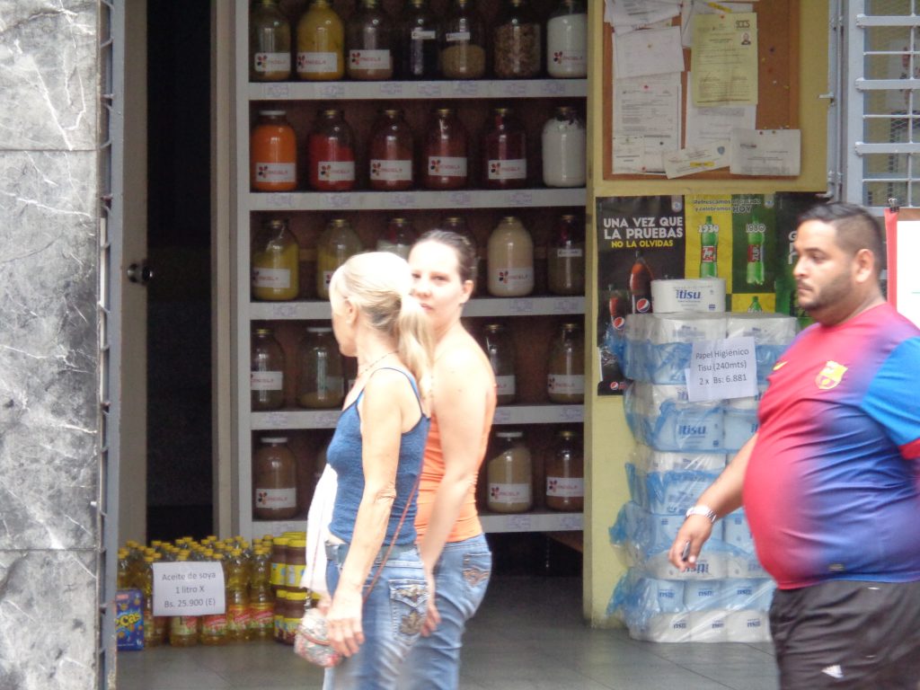 Зад минувачите се вижда асортиментът на това квартално магазинче в Каракас. Вдясно, зад едрия мъж, има купчина с тоалетна хартия по 6 881 боливара за две ролки. А долу вляво са стековете със соево олио–по 25 900 боливара за литър. Снимка: Къдринка Къдринова