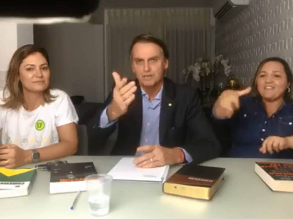 В нощта след победата си Болсонаро се обърна към бразилците по любимия си начин–през Фейсбук. До него беше младата му жена Мишел (в бяло) и интерпретаторка за хора с увреден слух, а на масата личаха Библията и конституцията, която новоизбраният президент се зарече да спазва. Снимка: Фейсбук 